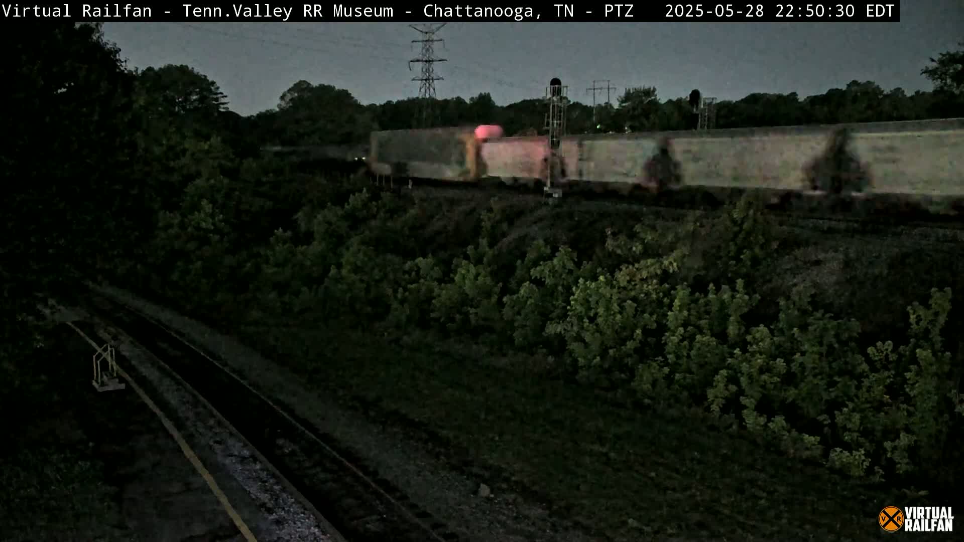 A long freight train moves through a dark, nighttime landscape beside a rail line bordered by trees.