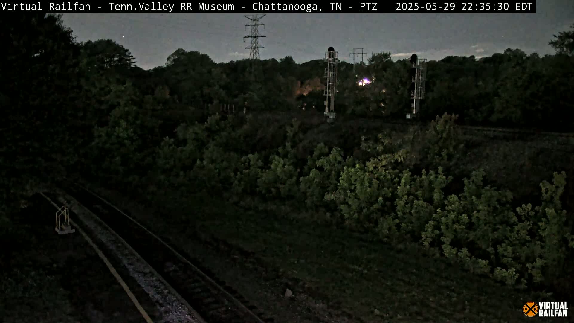 A nighttime, low-light view of train tracks running through a wooded area, with a tall structure and power lines visible in the background.