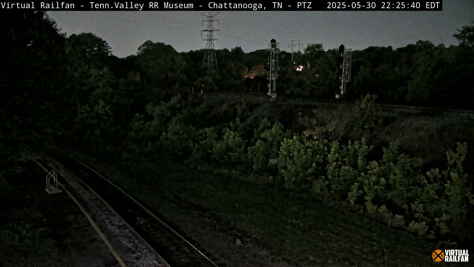 A nighttime, high-angle view shows a railway track surrounded by dark green vegetation, with tall electrical towers visible in the background under a twilight sky.