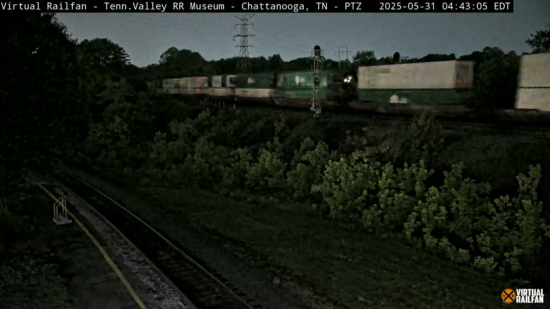 A long freight train passes by under a twilight sky, traveling along railroad tracks that are bordered by lush green vegetation.
