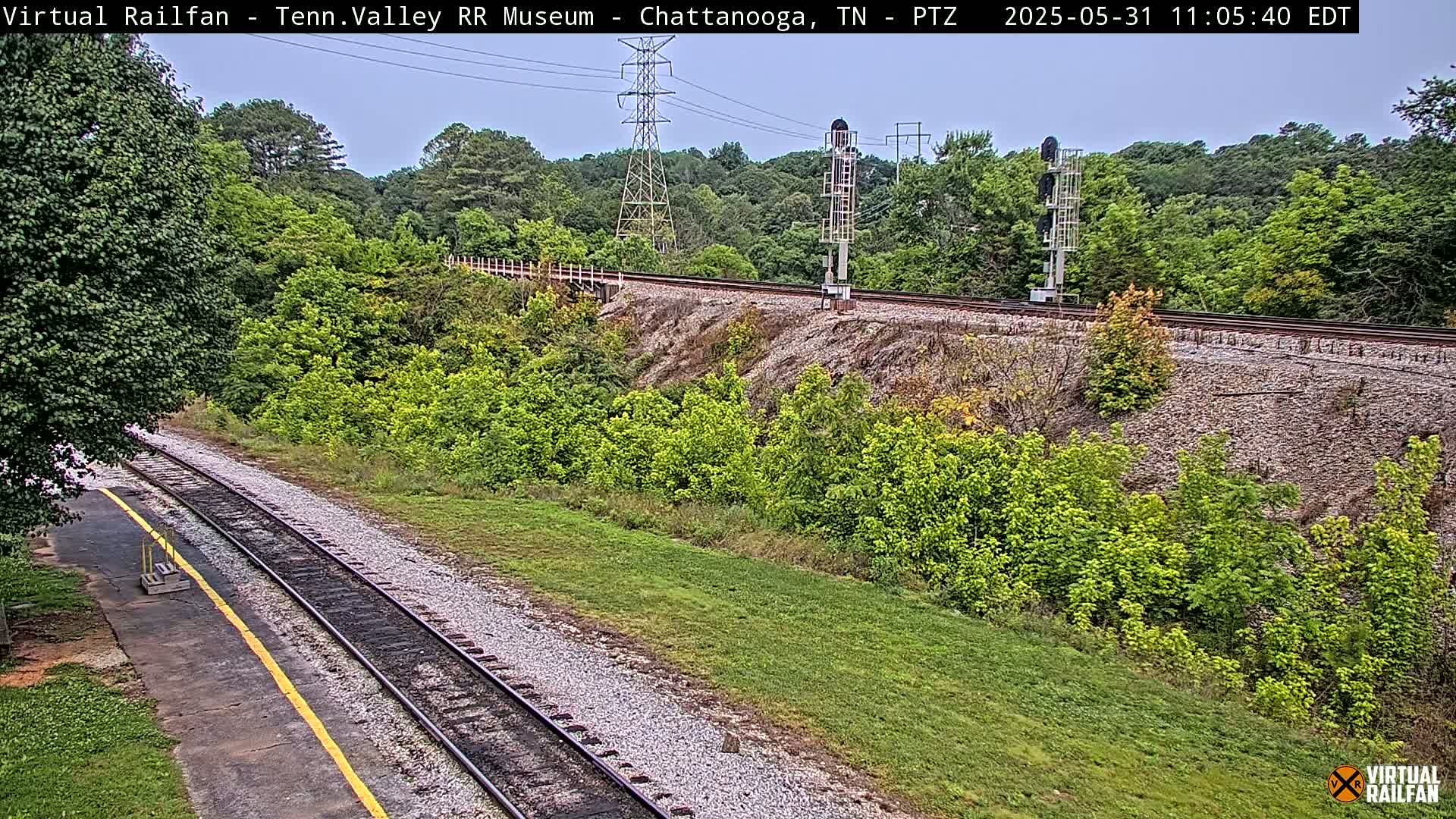 A mostly sunny day view of railroad tracks winding through a grassy area beside a lush green hillside with trees and power lines in the distance.