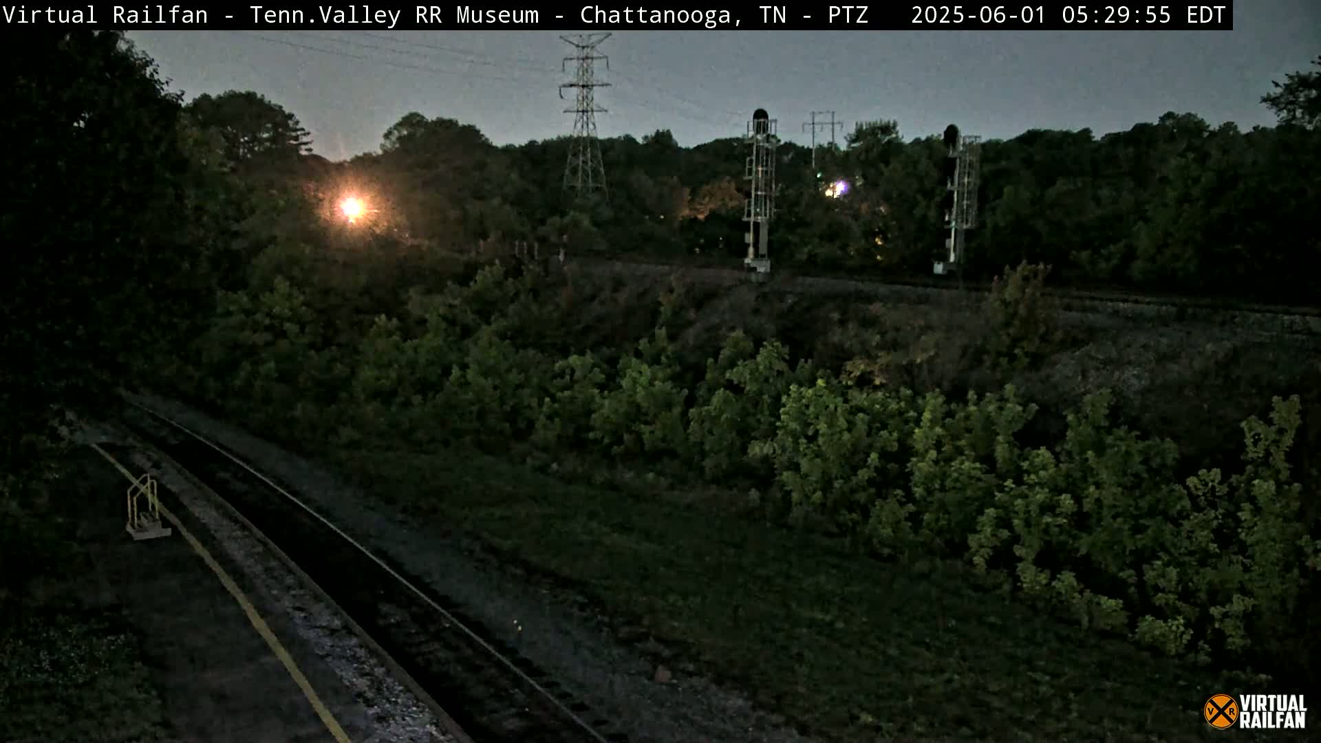 A twilight view of railroad tracks winding through a dark green, wooded area, with tall utility poles and signal structures visible in the distance.