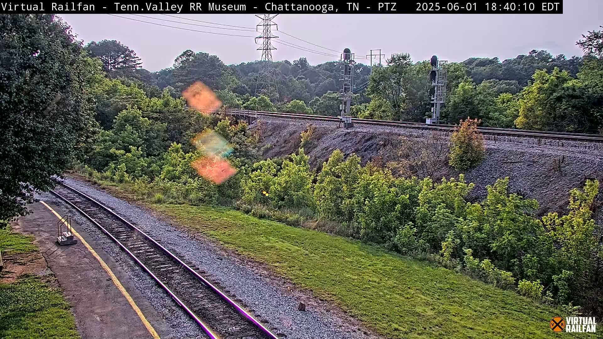 A view of railroad tracks winding through a grassy area and alongside a wooded hillside under an overcast sky.
