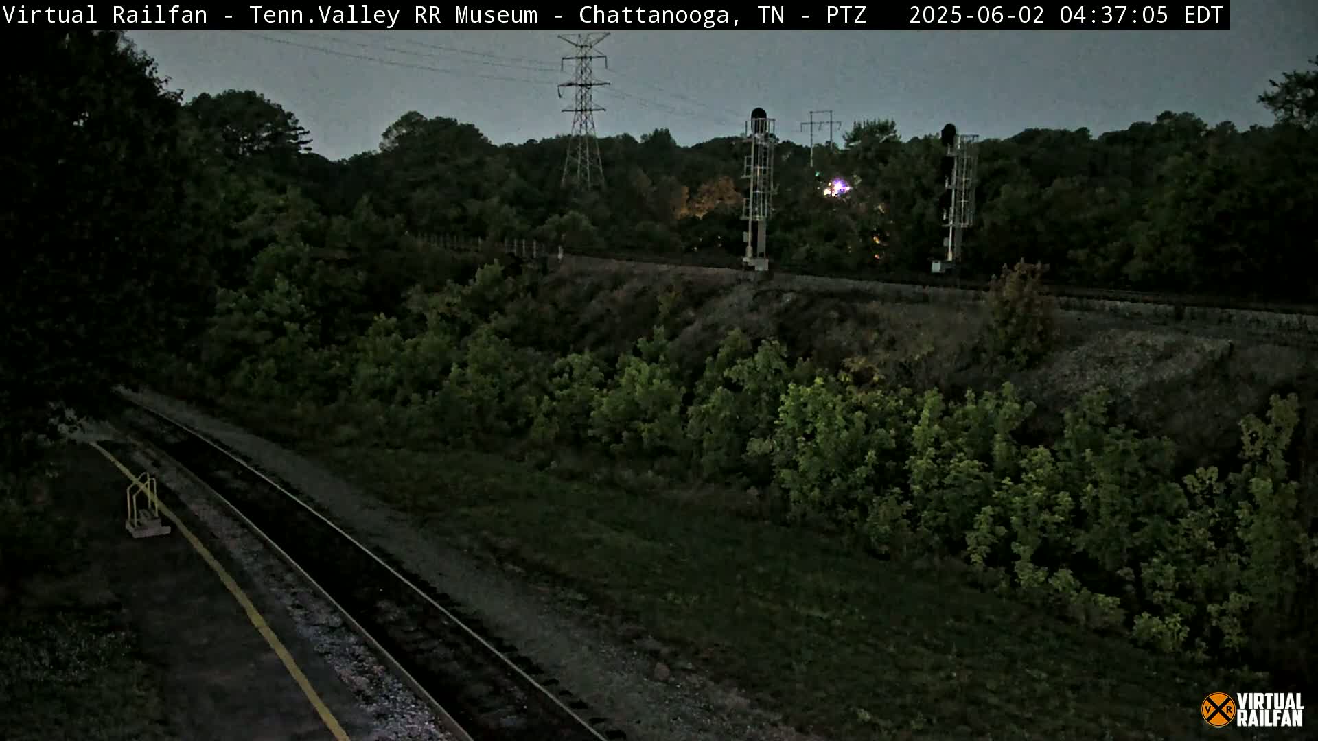 A dark, overcast evening view of a railroad track winding through a lush, green landscape, with power lines and signal equipment visible in the background.