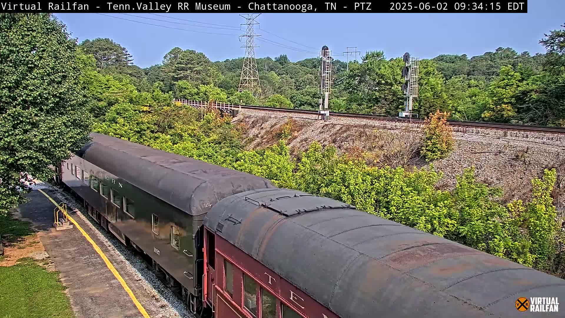 A long dark-colored passenger train sits on a track next to lush green vegetation on a sunny day.