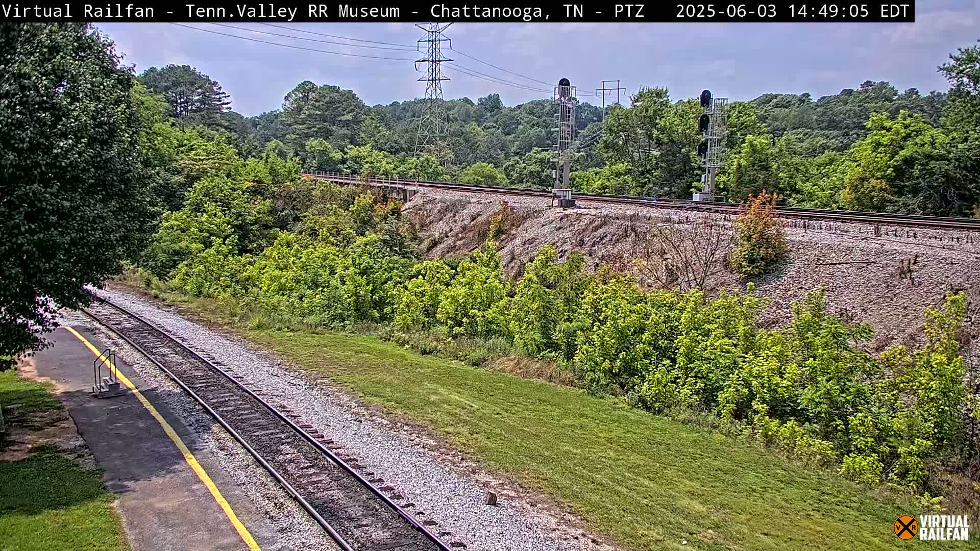 A mostly sunny view of a train track that curves to the right, surrounded by green vegetation and a small grassy area next to it.