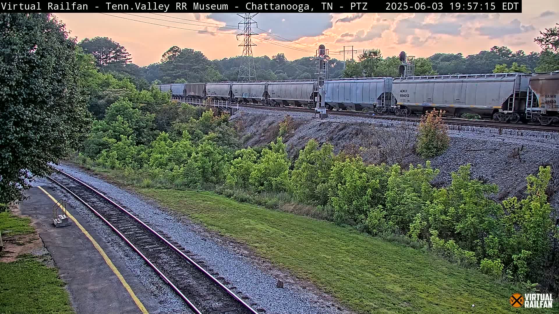 A long freight train passes by a grassy area next to railroad tracks at sunset.