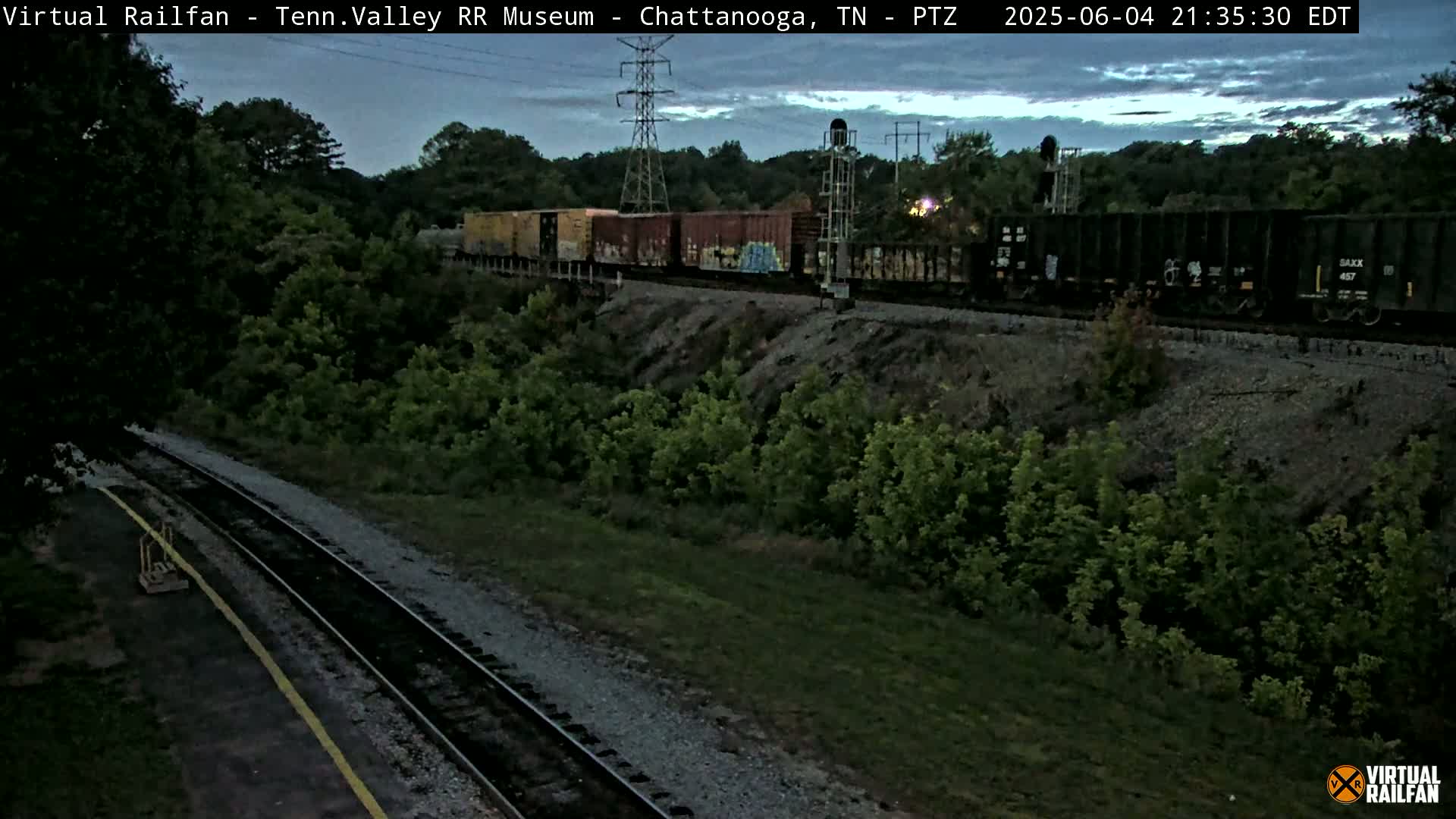 A freight train passes by under a twilight sky, traveling along railroad tracks bordered by green vegetation.