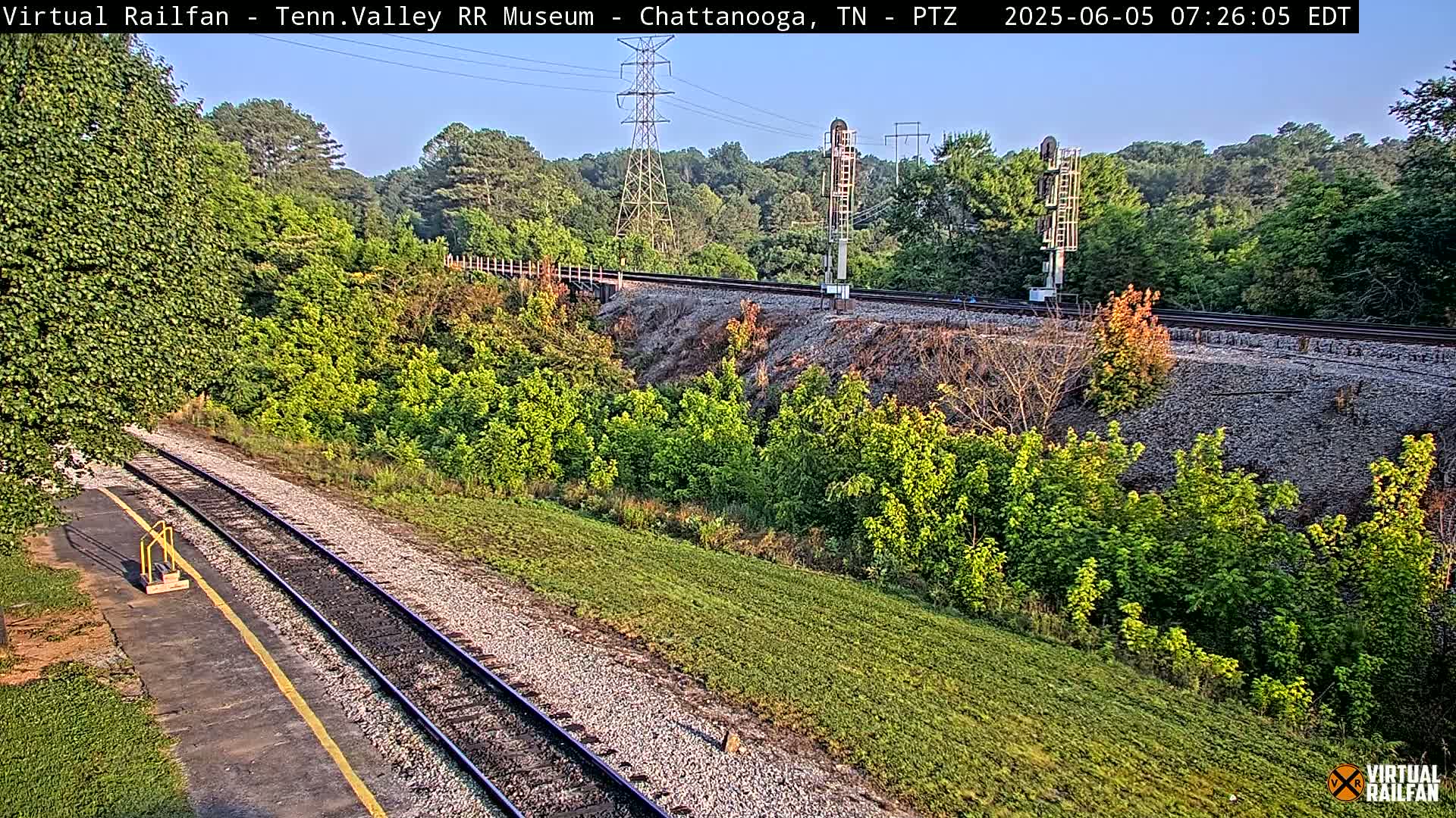 A section of train tracks runs through a grassy area, alongside a hillside covered in lush green vegetation, under a clear, bright sky.