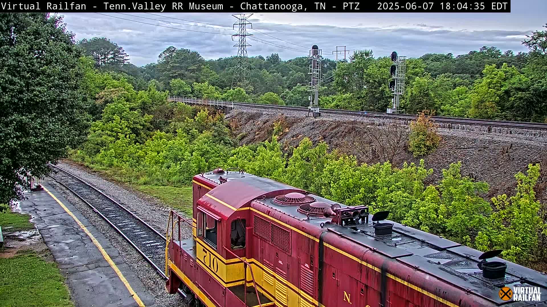 A maroon diesel locomotive sits on a rail line next to a lush green embankment under an overcast sky.