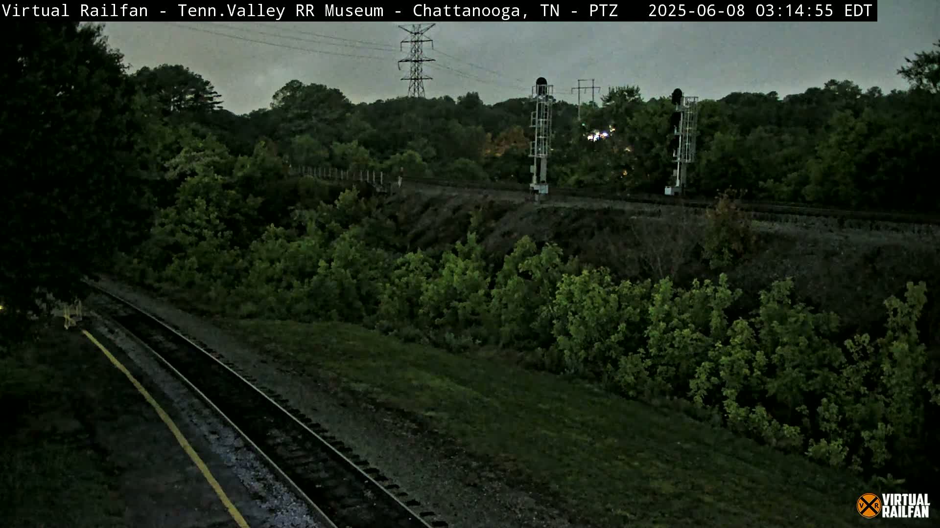 A railroad track cuts through a landscape of green trees and shrubs under an overcast sky.