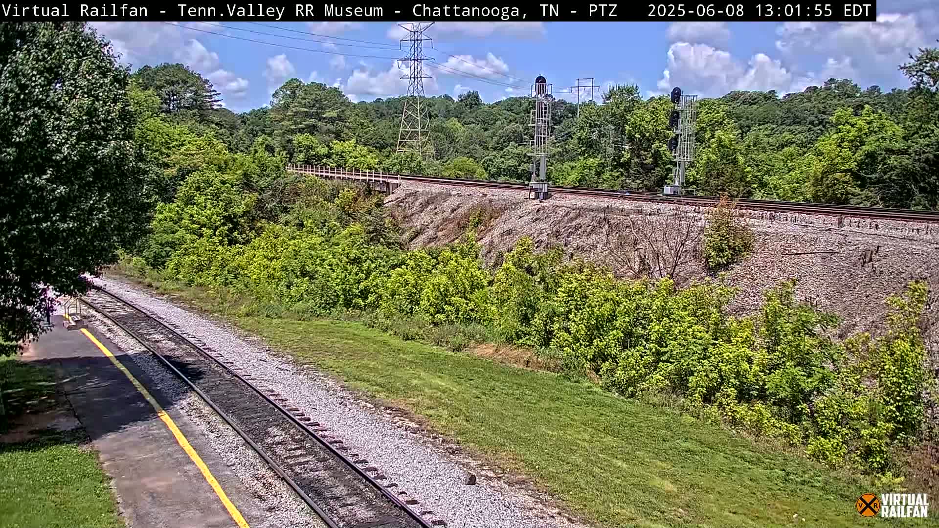 A partly sunny day reveals a train track winding through a lush green landscape, with a rocky embankment and trees in the background.