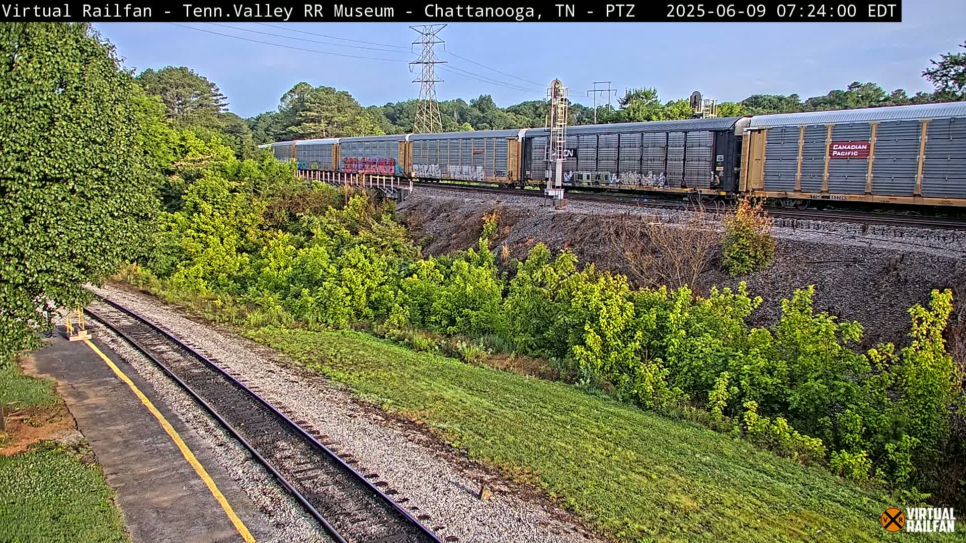 A long freight train passes by on a railway track next to lush green vegetation under a mostly clear sky.