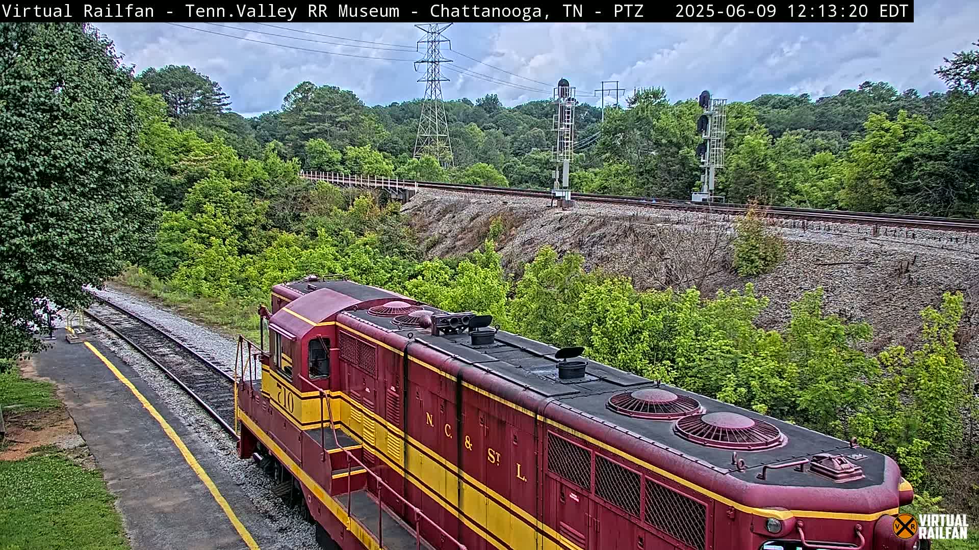 A red and yellow train is on a railroad track next to green trees and under a partly cloudy sky.