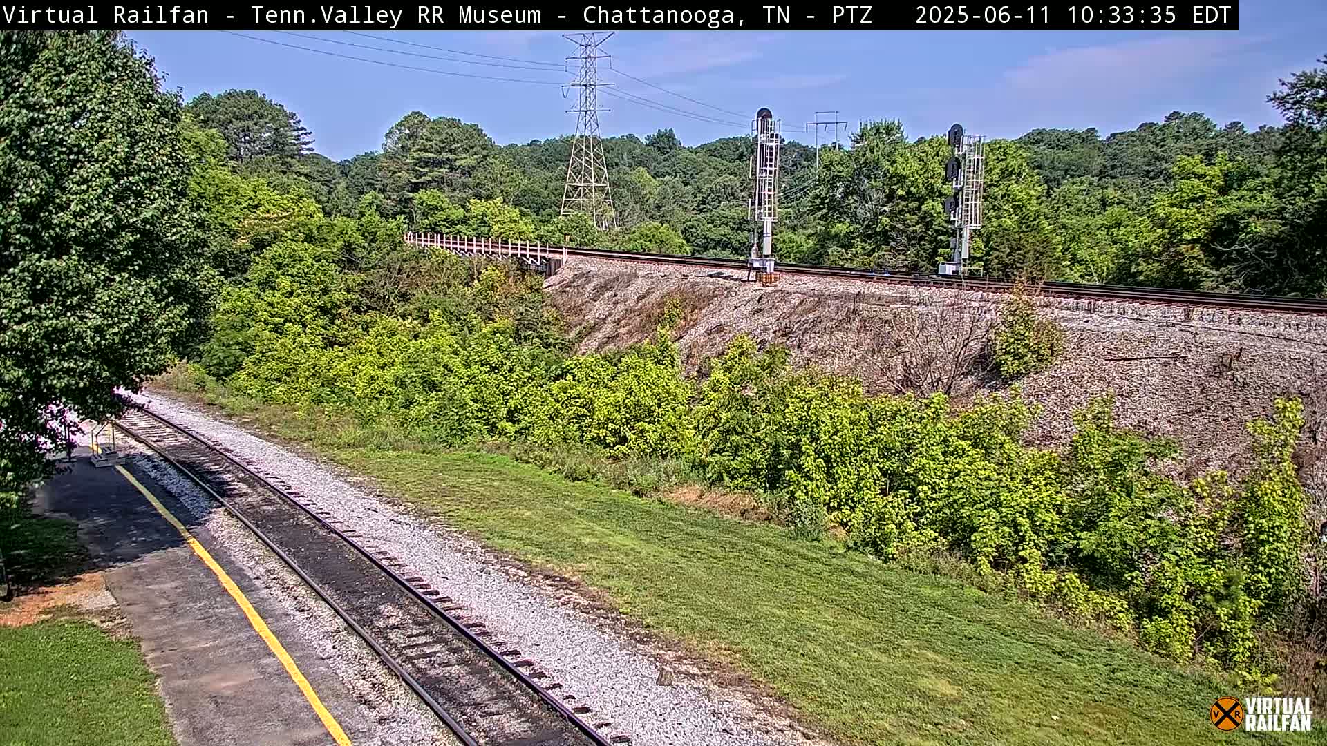 A high-angle view of a railway track winding through a grassy area, bordered by lush green vegetation and trees under a mostly clear sky.