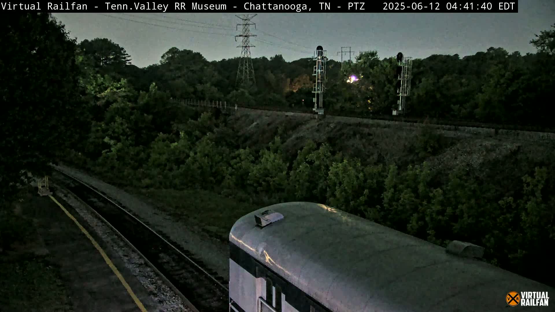 A silver train car is visible on a set of train tracks at dusk, surrounded by lush green vegetation and power lines.