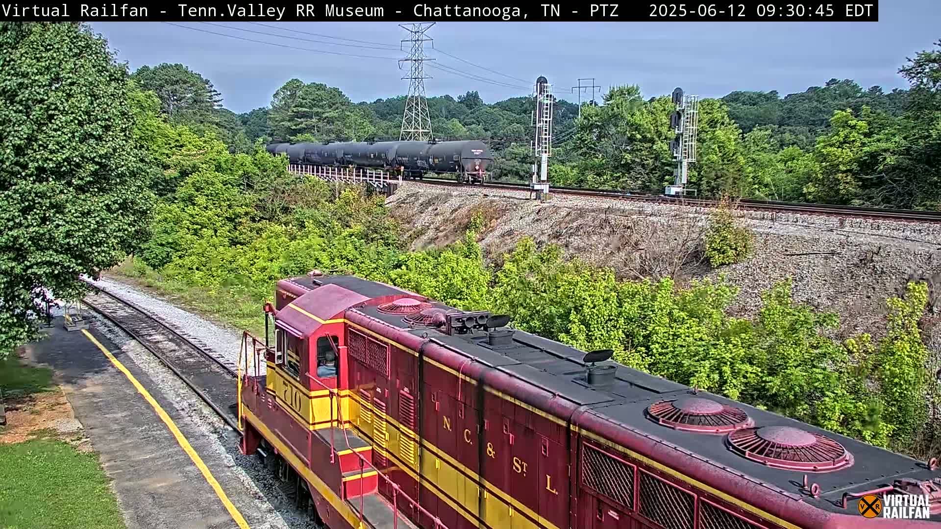 A red and yellow diesel locomotive sits on a railway track next to a lush green embankment, with a long freight train visible in the distance under a partly cloudy sky.
