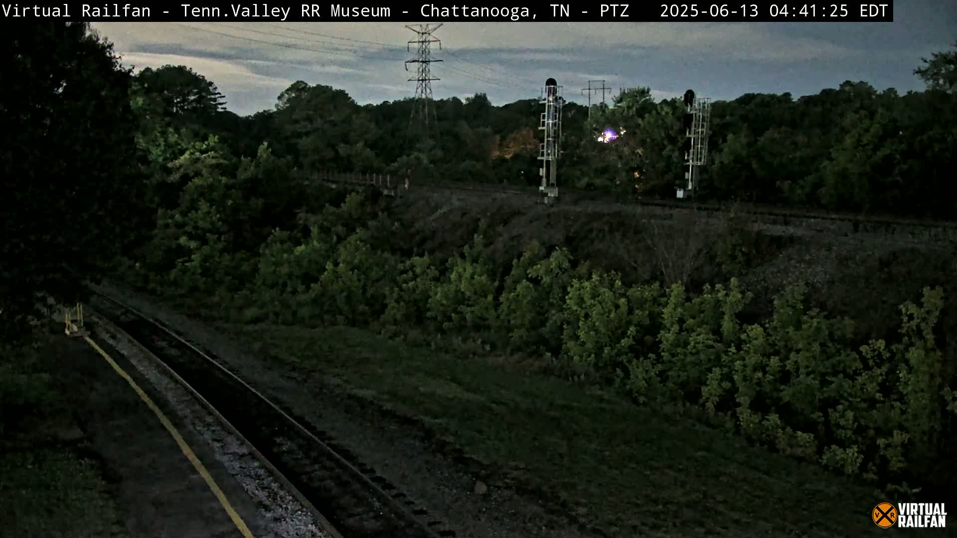 A twilight view of railroad tracks winding through a lush green landscape, with a tall transmission tower and signal lights visible in the background.
