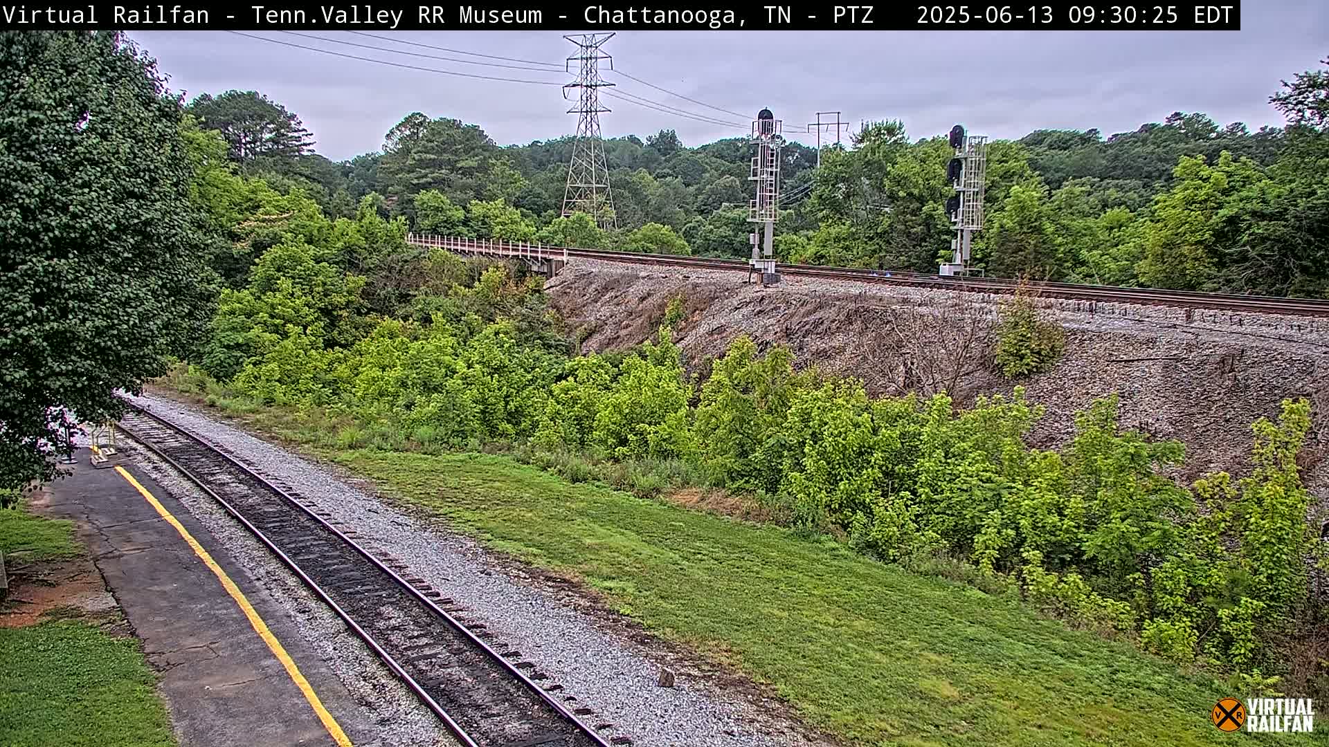 A mostly cloudy day shows a railway track running through a grassy area next to lush green trees and shrubs, with a tall transmission tower and signal in the distance.