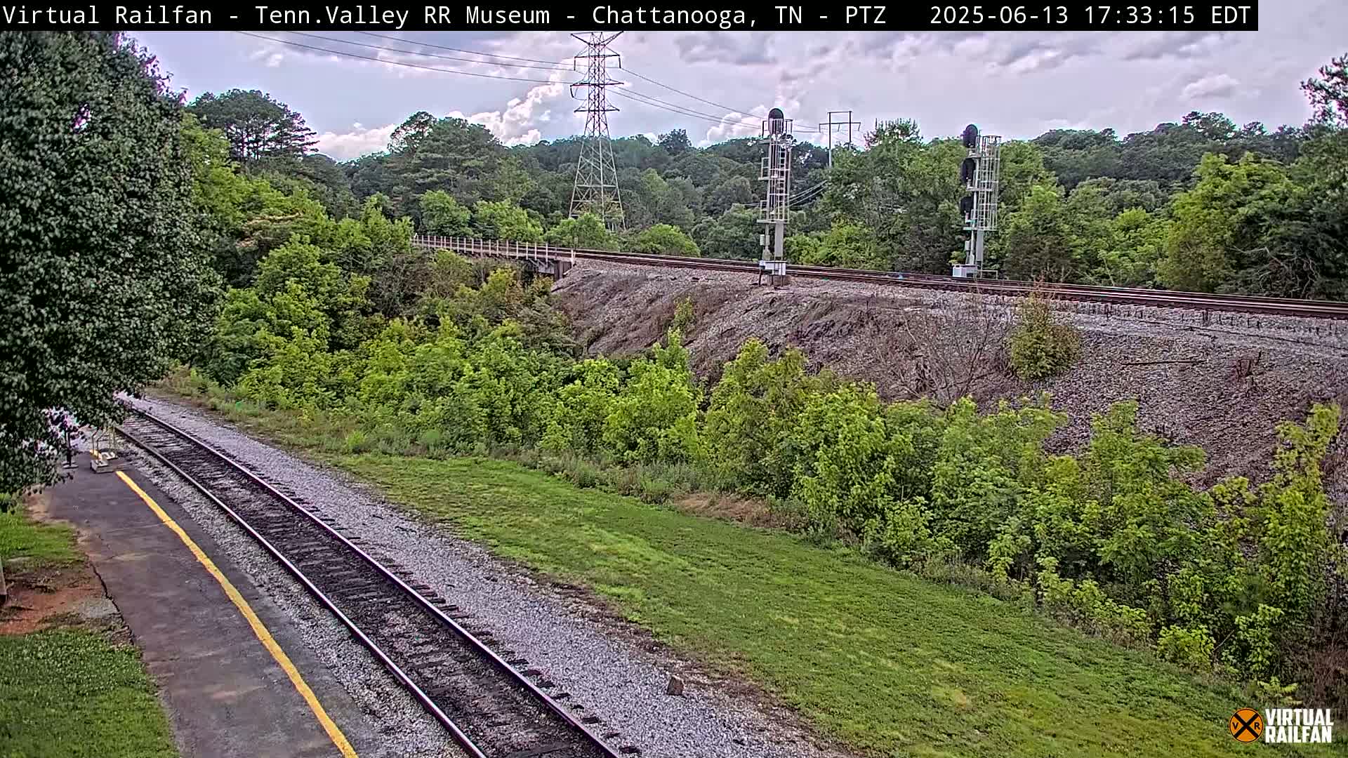 A partly cloudy day reveals a view of a railroad track curving through a lush green landscape, with a tall transmission tower and signal lights visible in the distance.