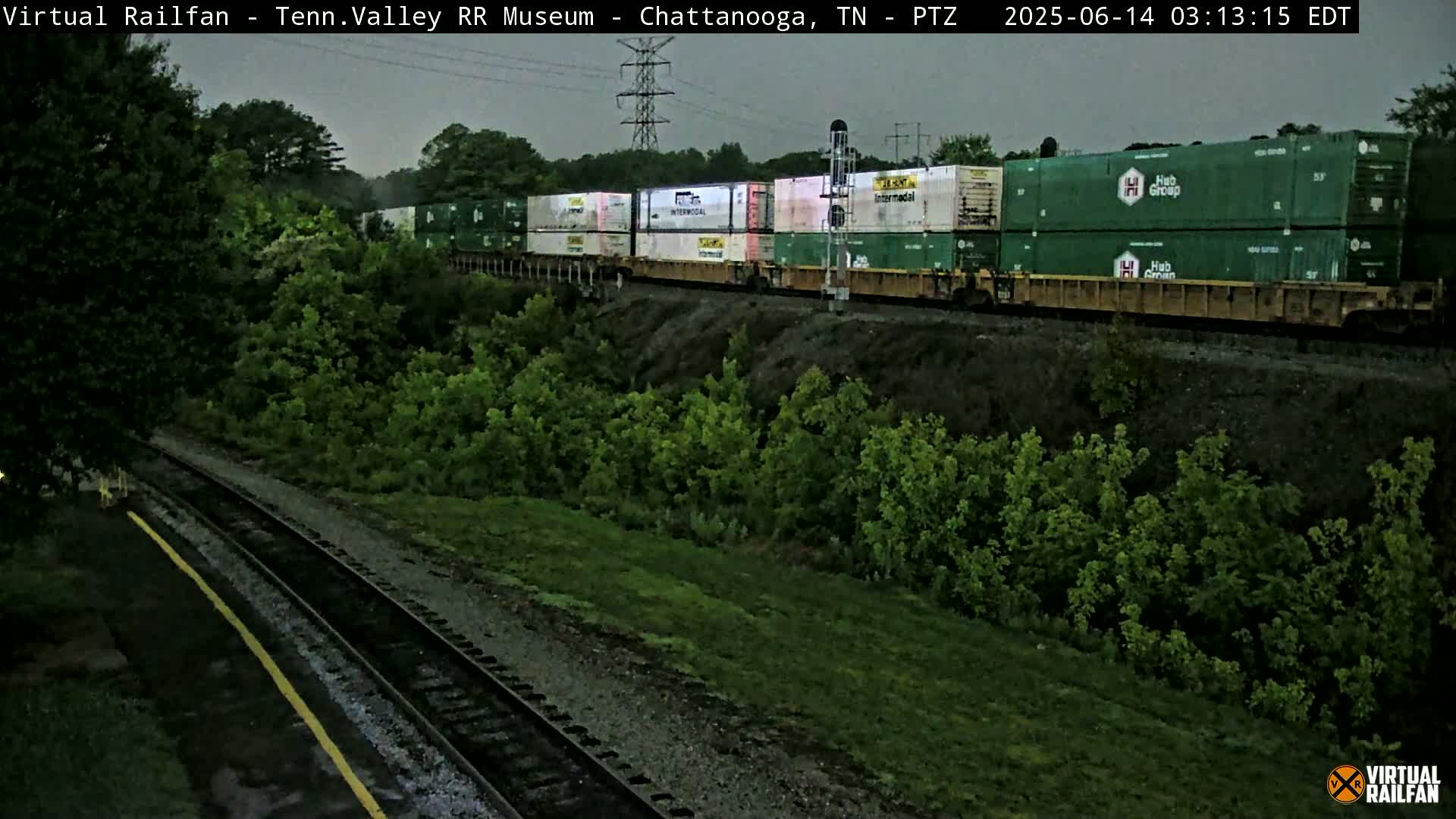 A freight train carrying green and white shipping containers passes by lush green vegetation under an overcast sky.