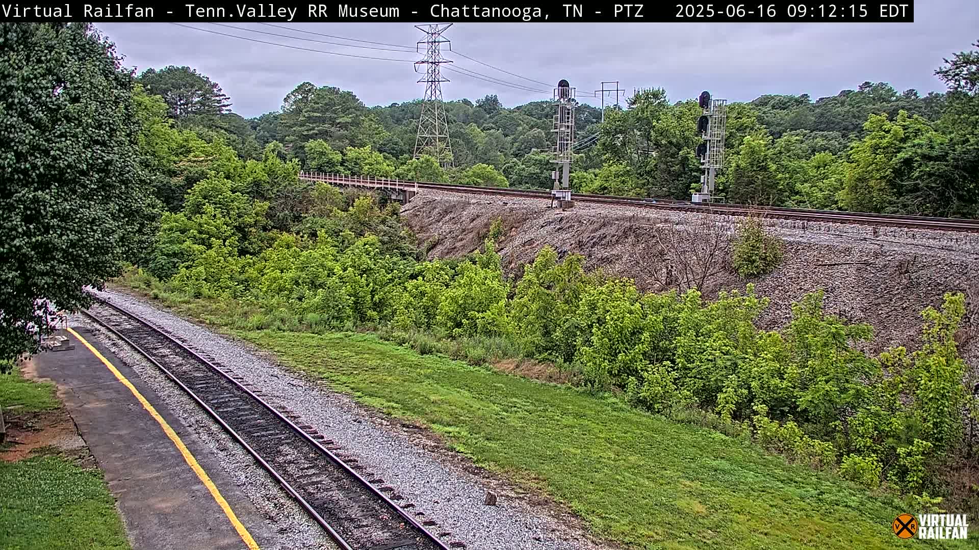 Railroad tracks extend from a grassy area alongside a tree-lined embankment under an overcast sky.