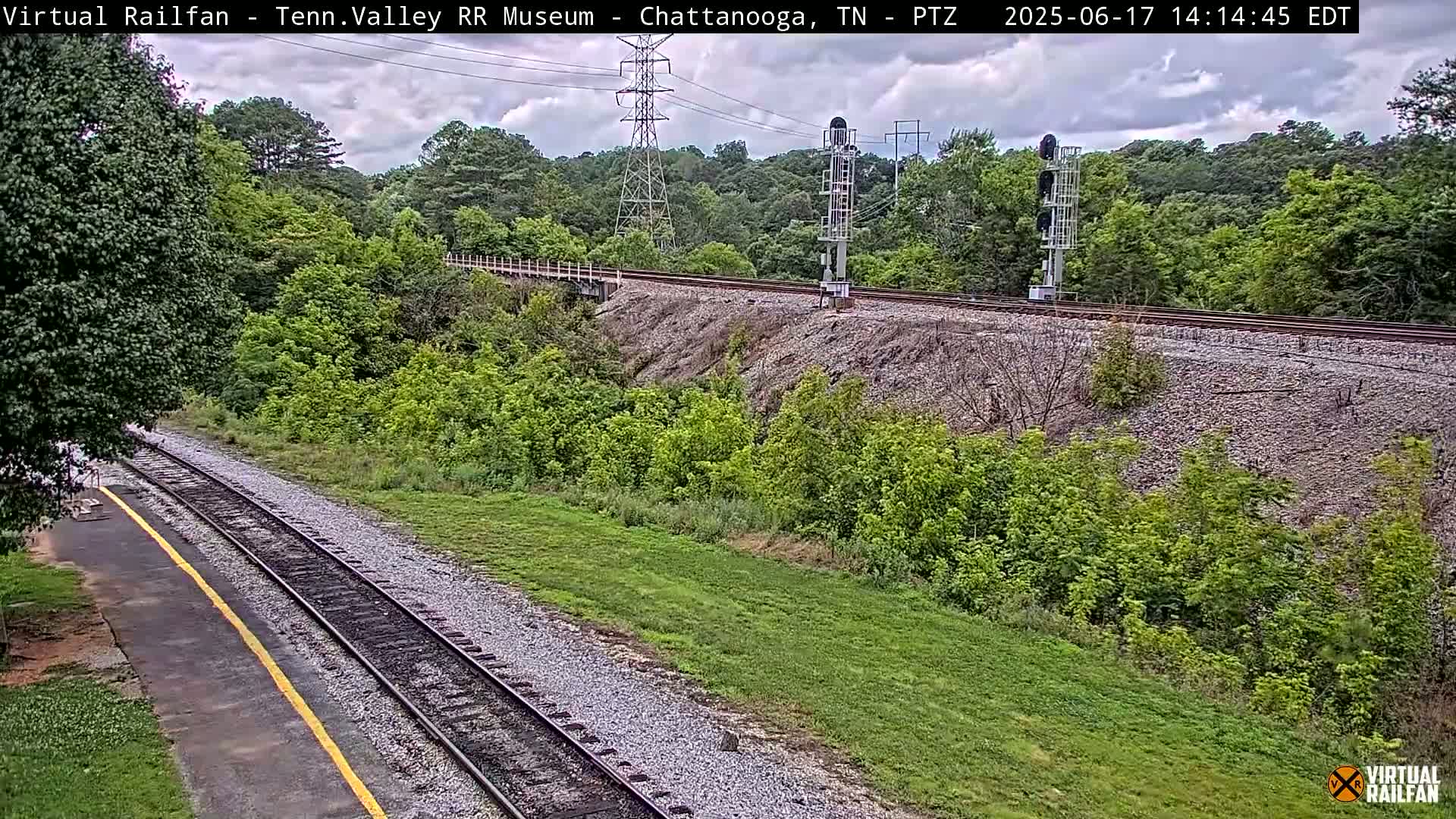 A mostly sunny day shows a railway track running through a verdant landscape, with trees and grass alongside a sloped embankment.