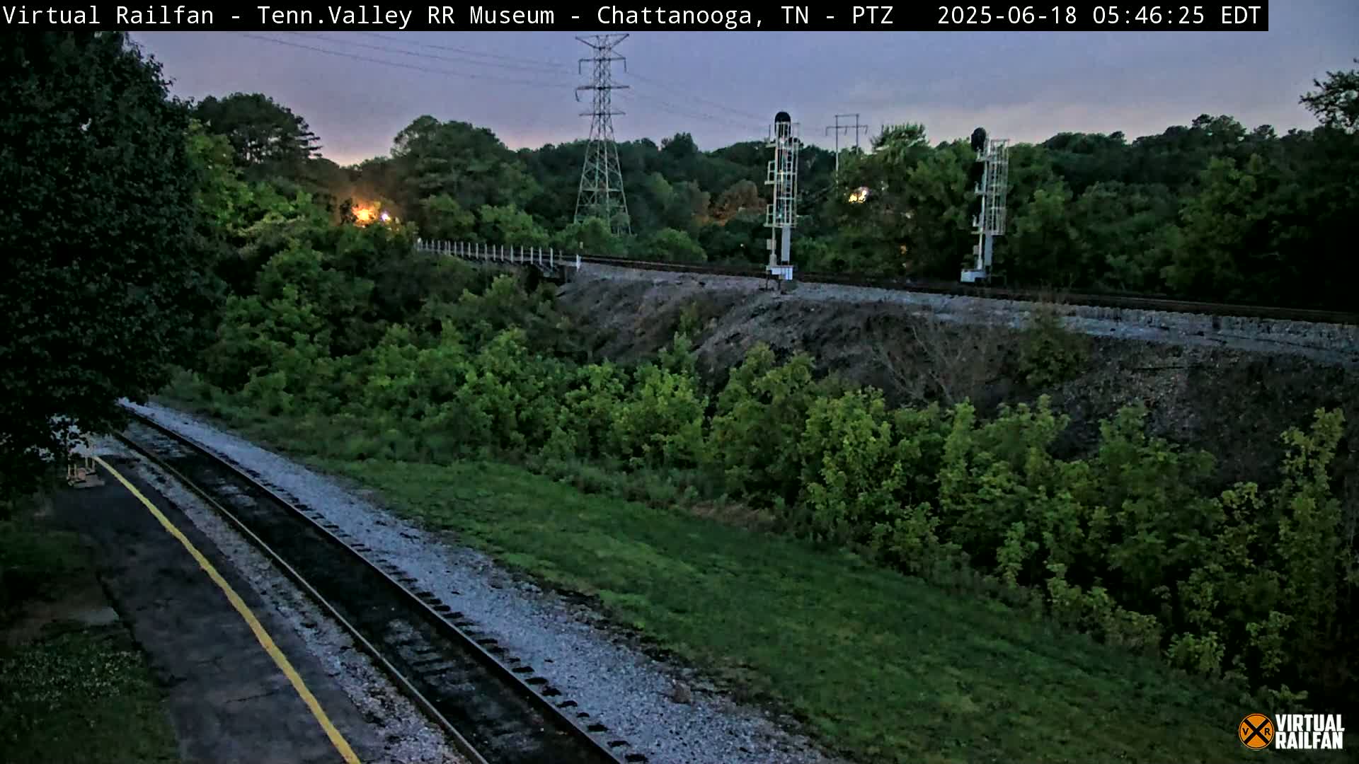 A twilight view of train tracks winding through a lush green landscape, with a small bridge visible in the distance.