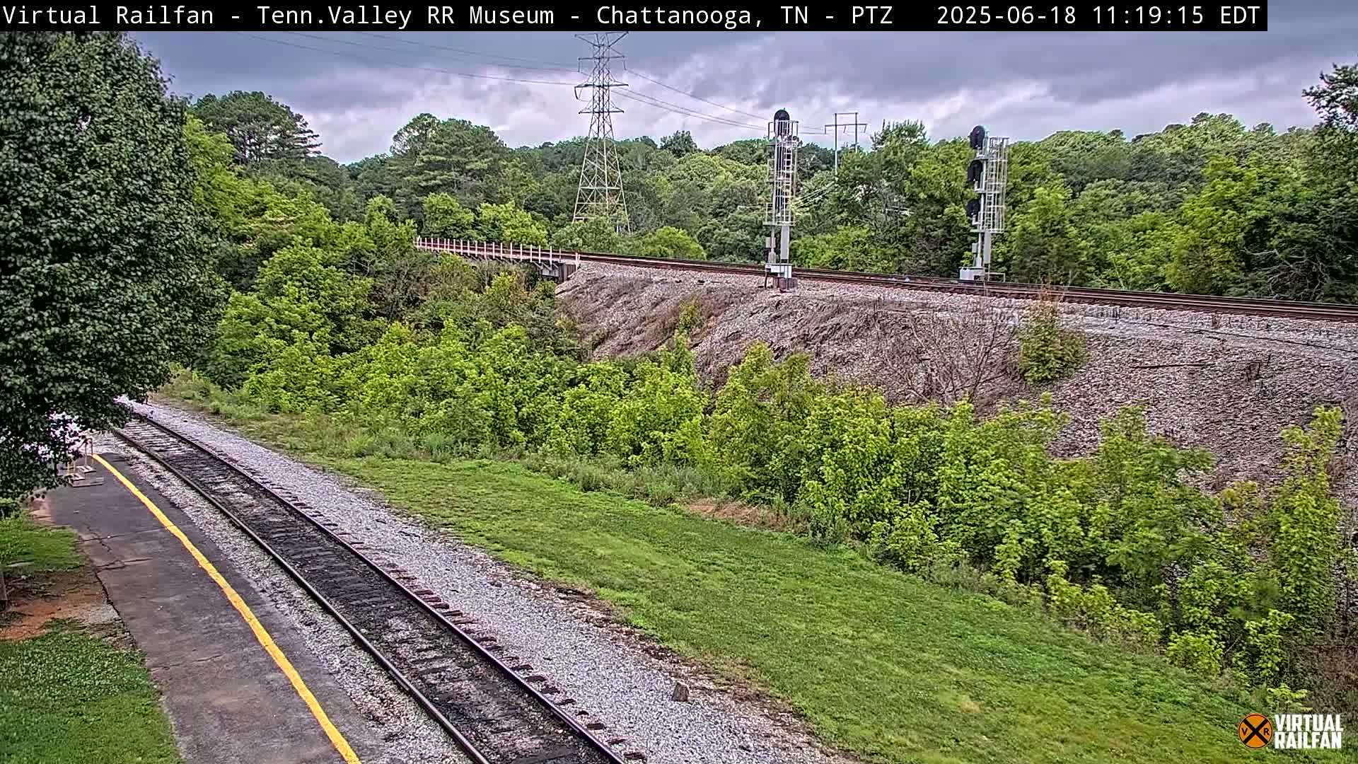 A railroad track runs through a lush green landscape under a partly cloudy sky.