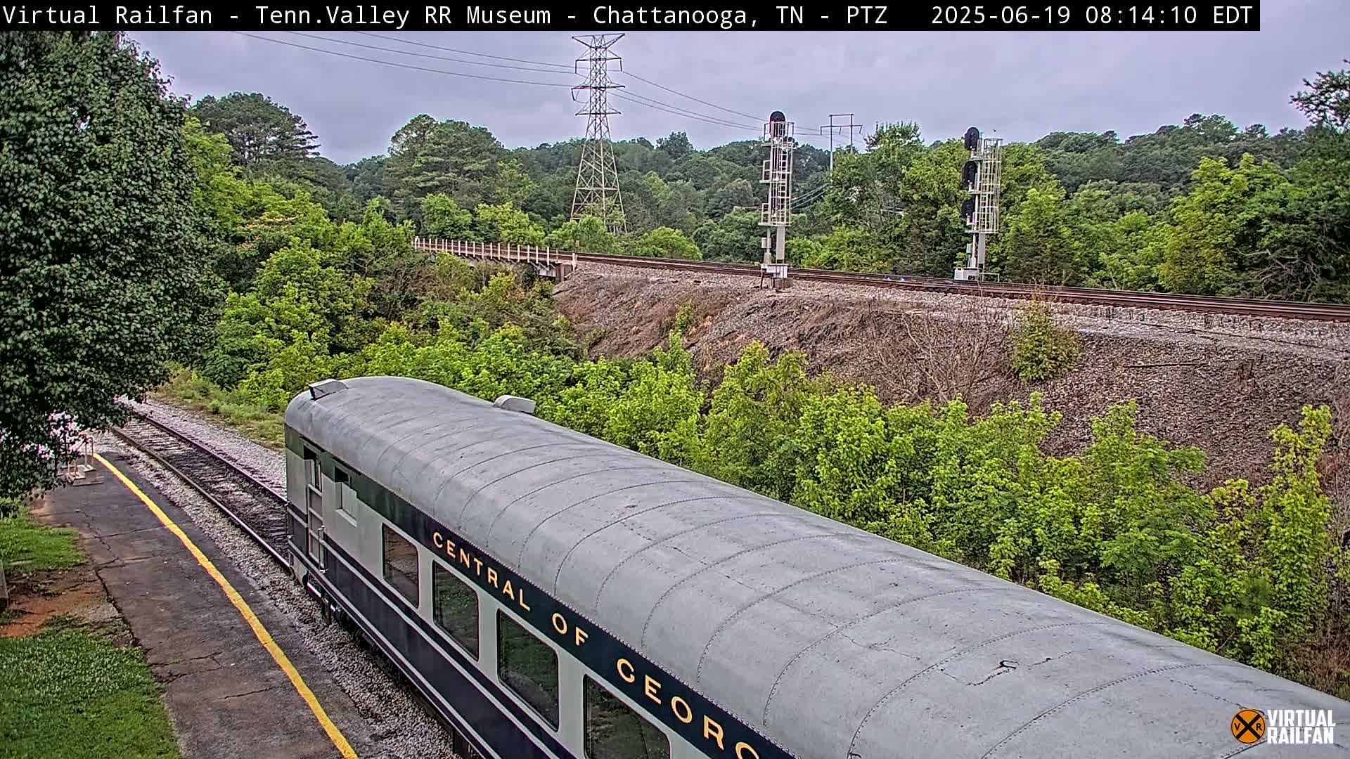 A gray passenger train car, labeled "Central of Geor[gia]", is visible on railroad tracks next to lush green vegetation under an overcast sky.