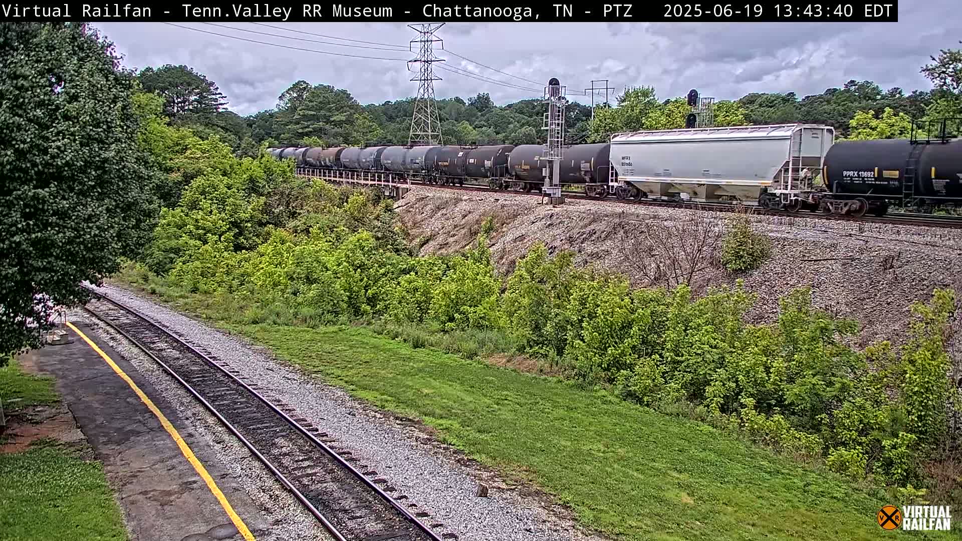 A long freight train passes by on a railway track next to a grassy area under partly cloudy skies.