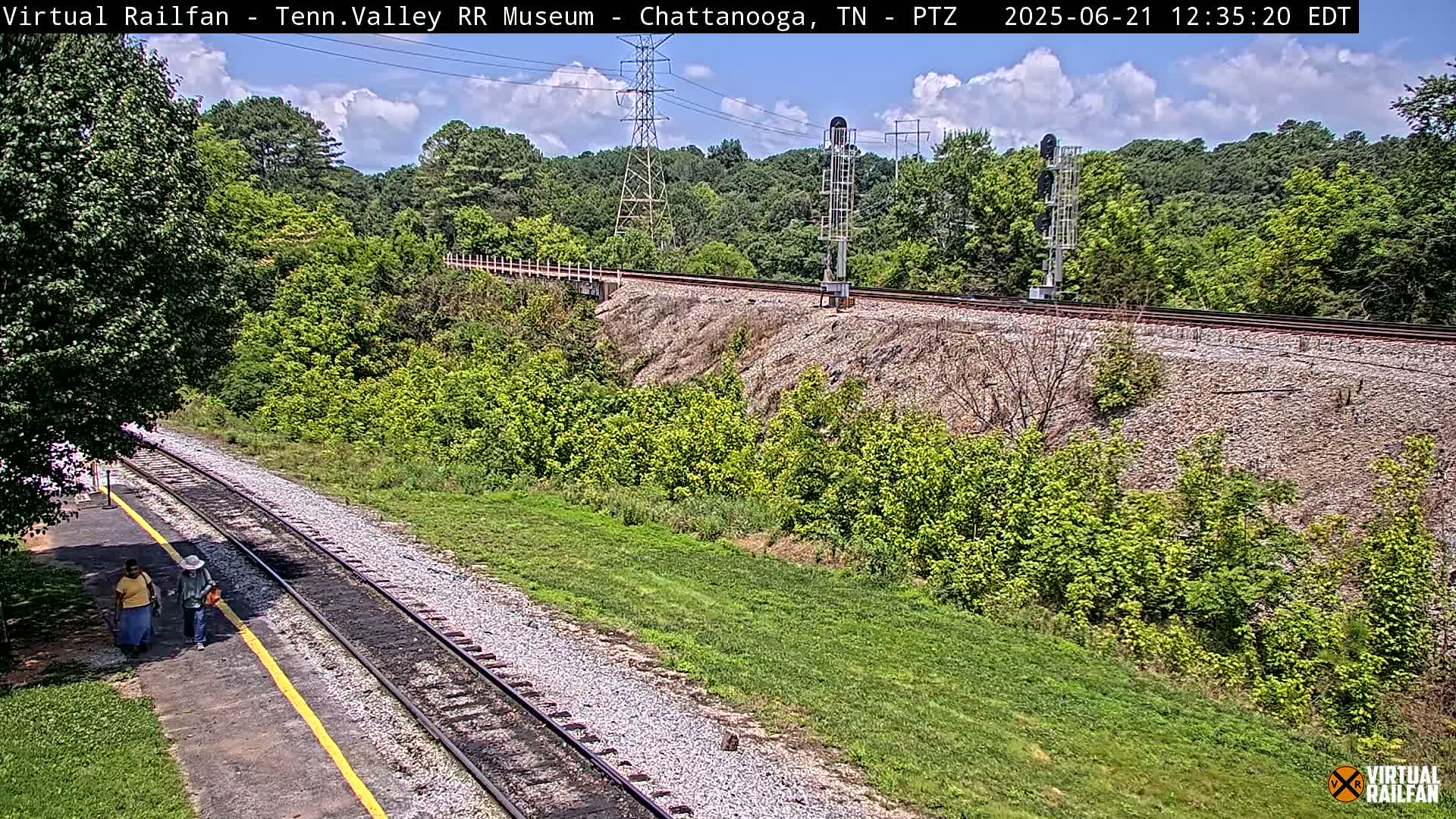 A partly cloudy day shows a railway track running through a lush green landscape, with two people walking along a path beside the tracks.