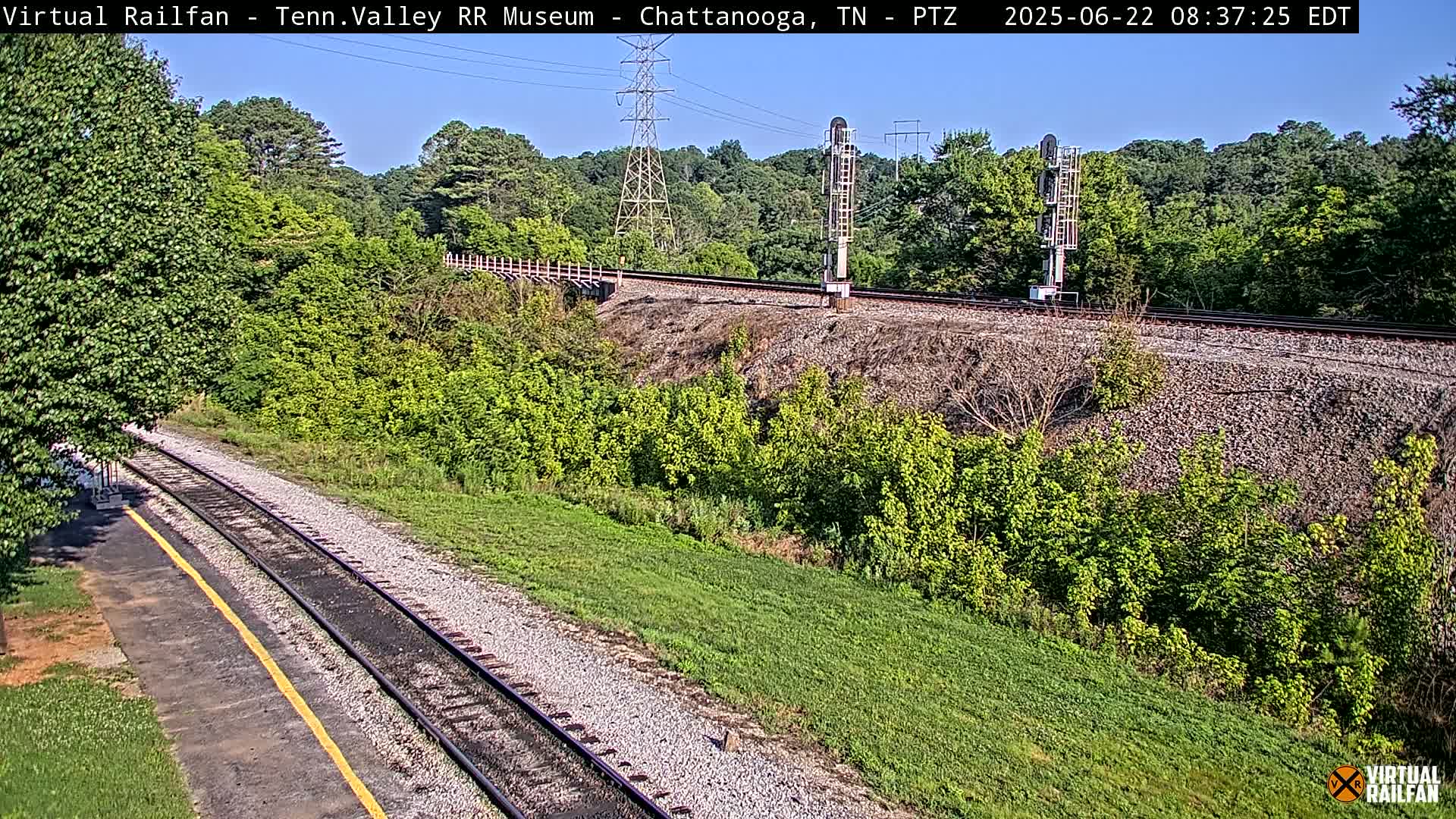 A sunny day view of railroad tracks that run alongside a grassy area with trees and bushes, passing under a power line.