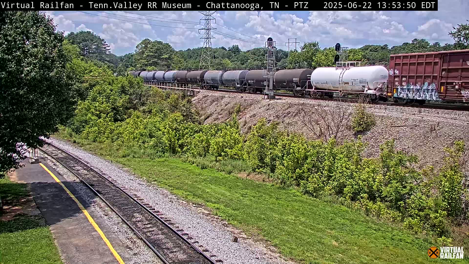 A long freight train passes by a grassy area next to train tracks on a partly sunny day.