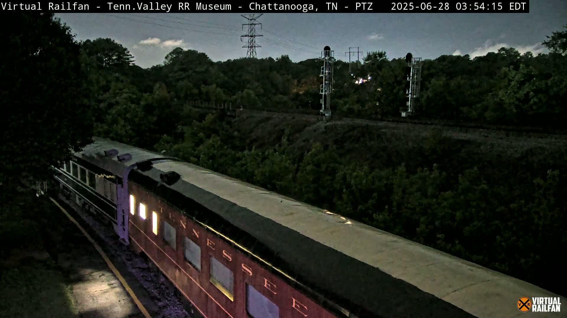 A passenger train is seen at night, under a mostly cloudy sky, alongside a treeline and railway infrastructure.