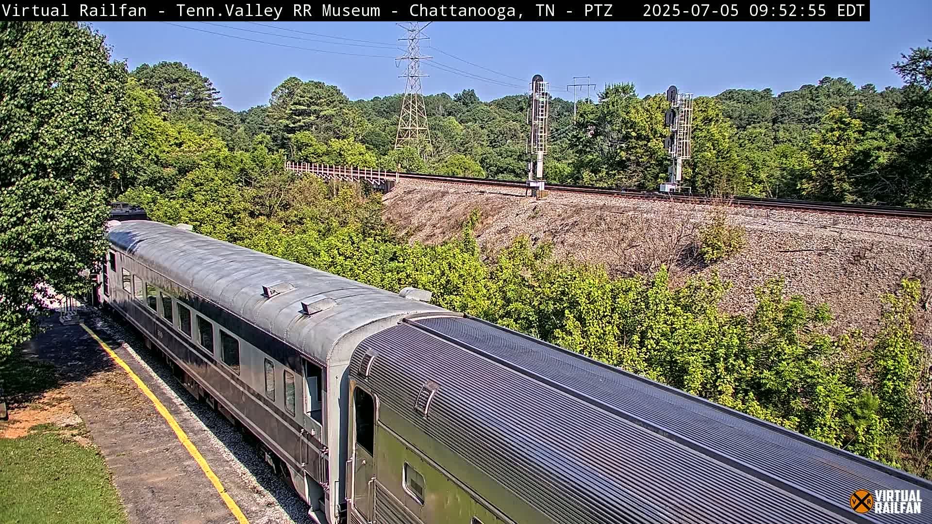 A long, gray passenger train car sits on a railway track next to a lush green hillside on a sunny day.