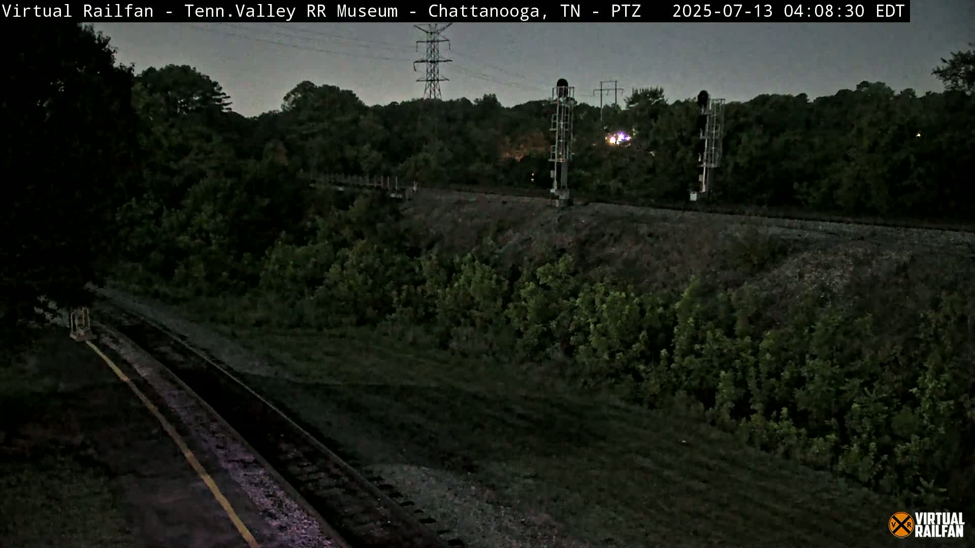 A nighttime view of railroad tracks bordered by lush green vegetation under a dusky sky.