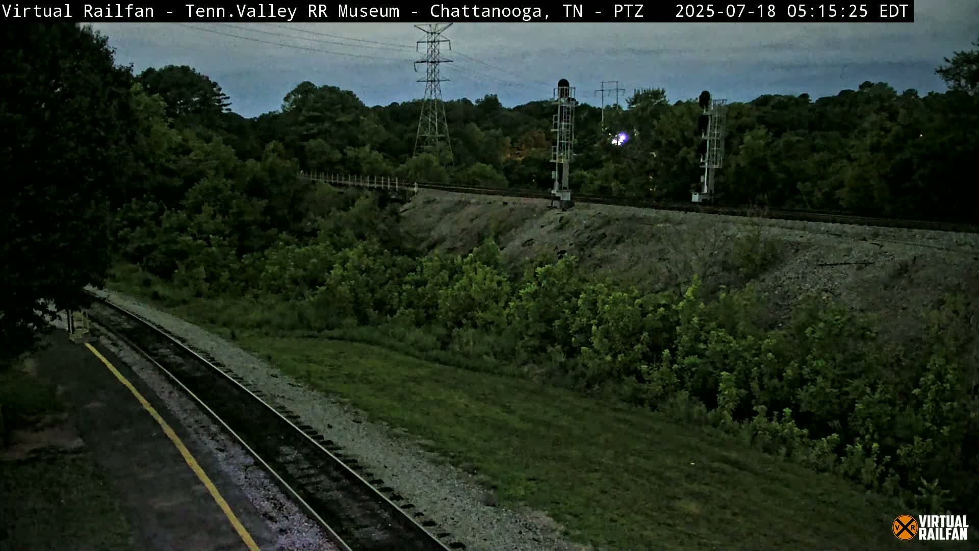 A high-angle, twilight view shows a railroad track winding through a lush green landscape, with power lines and signal equipment visible.