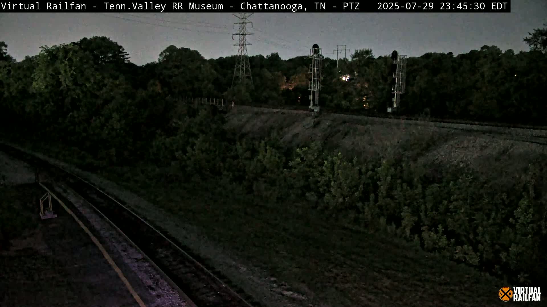 A high-angle, twilight view shows a railroad track winding through a lush green landscape, with power lines and signal equipment visible.