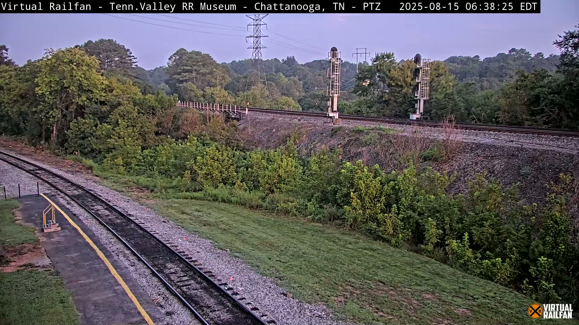A high-angle, twilight view shows a railroad track winding through a lush green landscape, with power lines and signal equipment visible.
