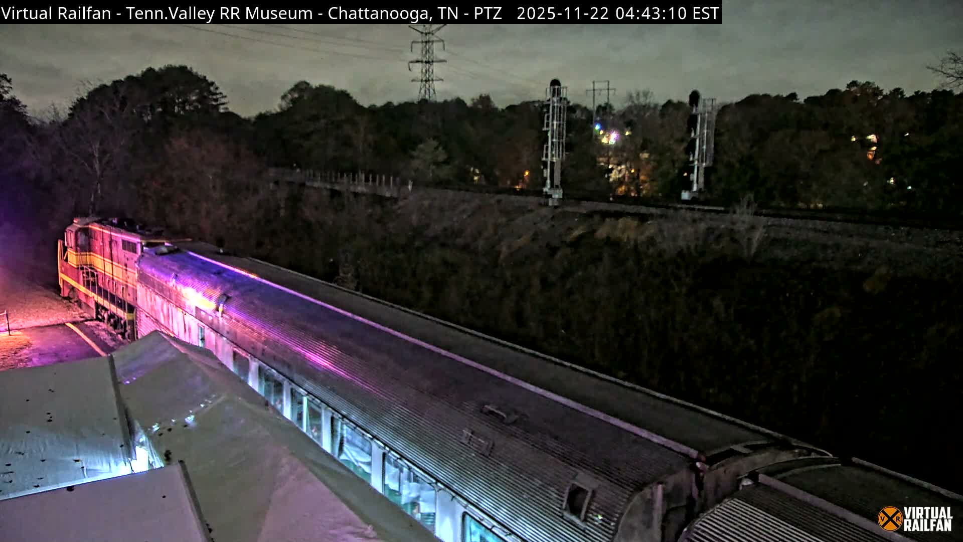 A long train, featuring an orange and yellow locomotive and passenger cars illuminated with vibrant purple light, is parked alongside a large roof structure on an overcast night.