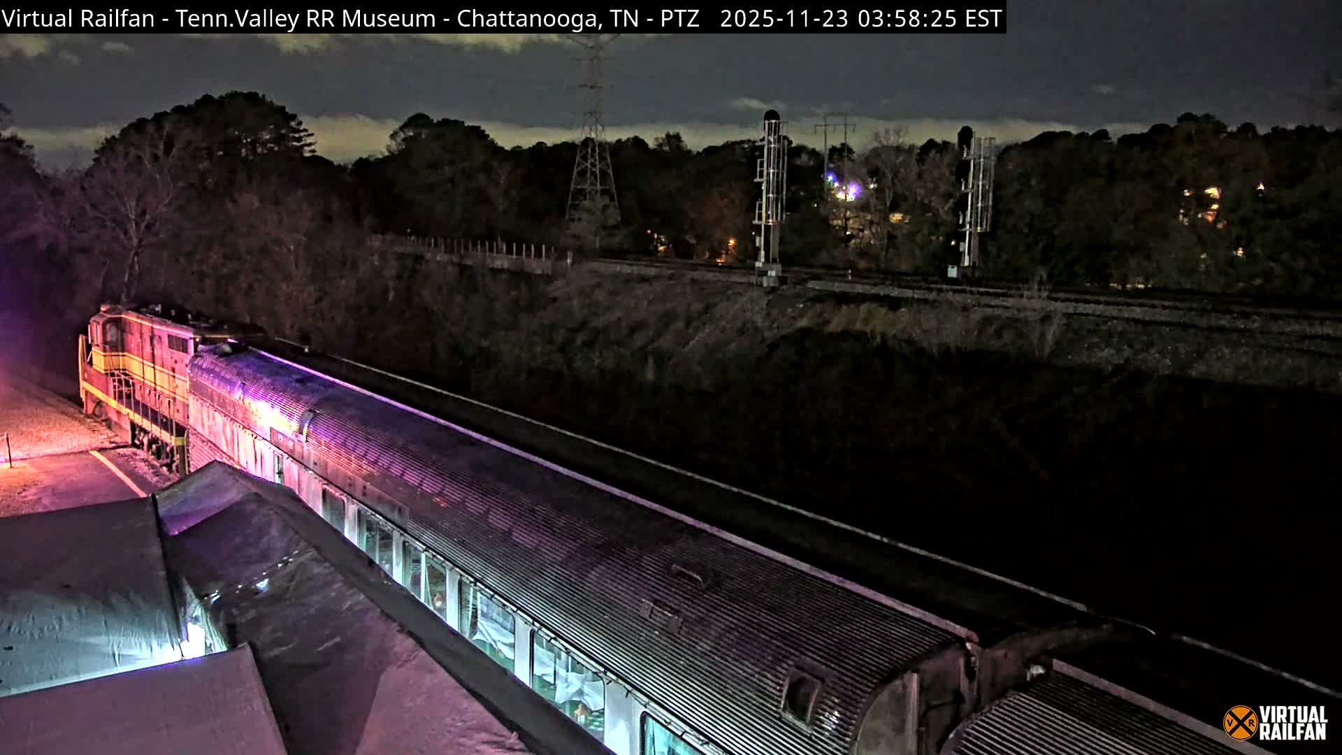 An elevated view at night shows a parked train with its cars illuminated in purplish light, alongside a platform, with additional train tracks and forested hills visible in the background under an overcast sky.