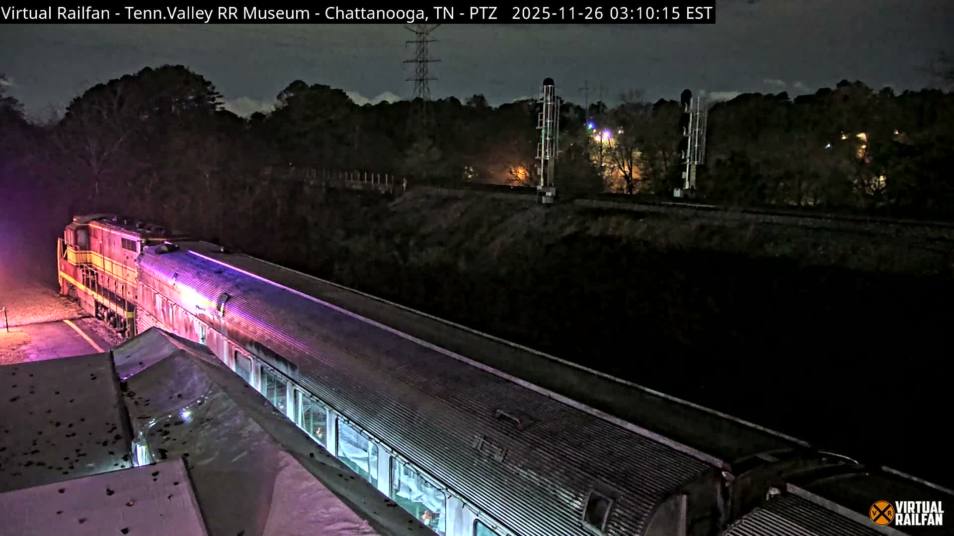 A vibrant, colorfully lit train, consisting of a red and yellow locomotive and silver passenger cars, is parked outdoors at night under an overcast sky, with railway signals visible in the distance.