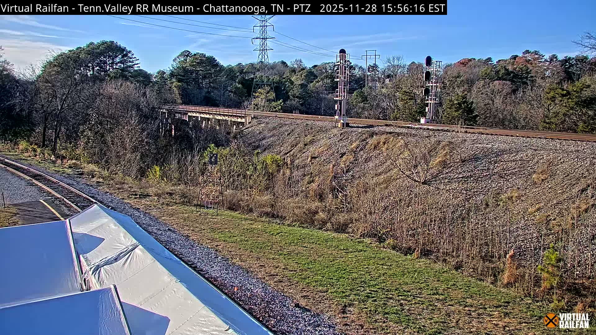 A sunny, clear day reveals a railroad scene featuring multiple tracks, a distant bridge, a tall signal pole, and a steep gravel embankment, all set against a backdrop of trees and a bright blue sky.