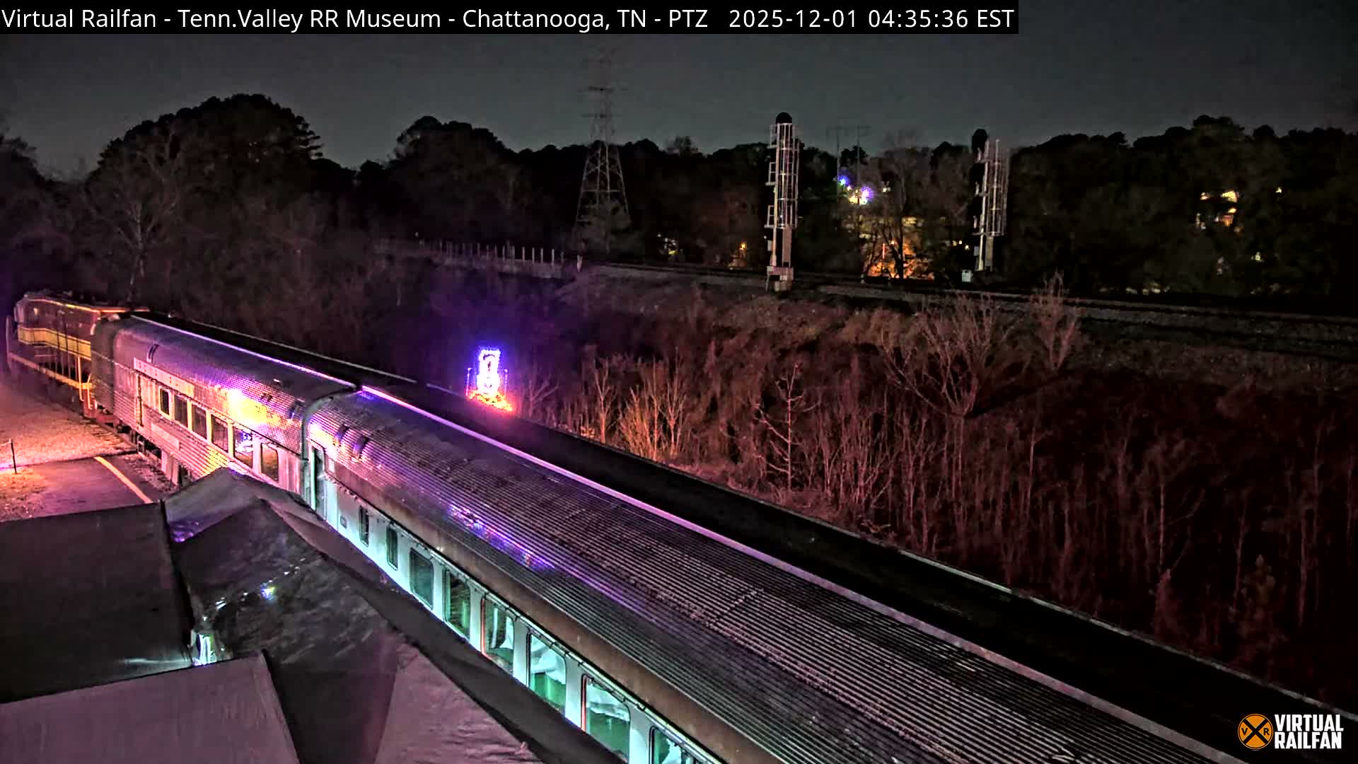 A train with brightly lit passenger cars, illuminated in purple, pink, and green hues, is parked next to a covered platform at night with clear weather, while in the background, bare trees line a hillside beyond which additional tracks and distant lights are visible.