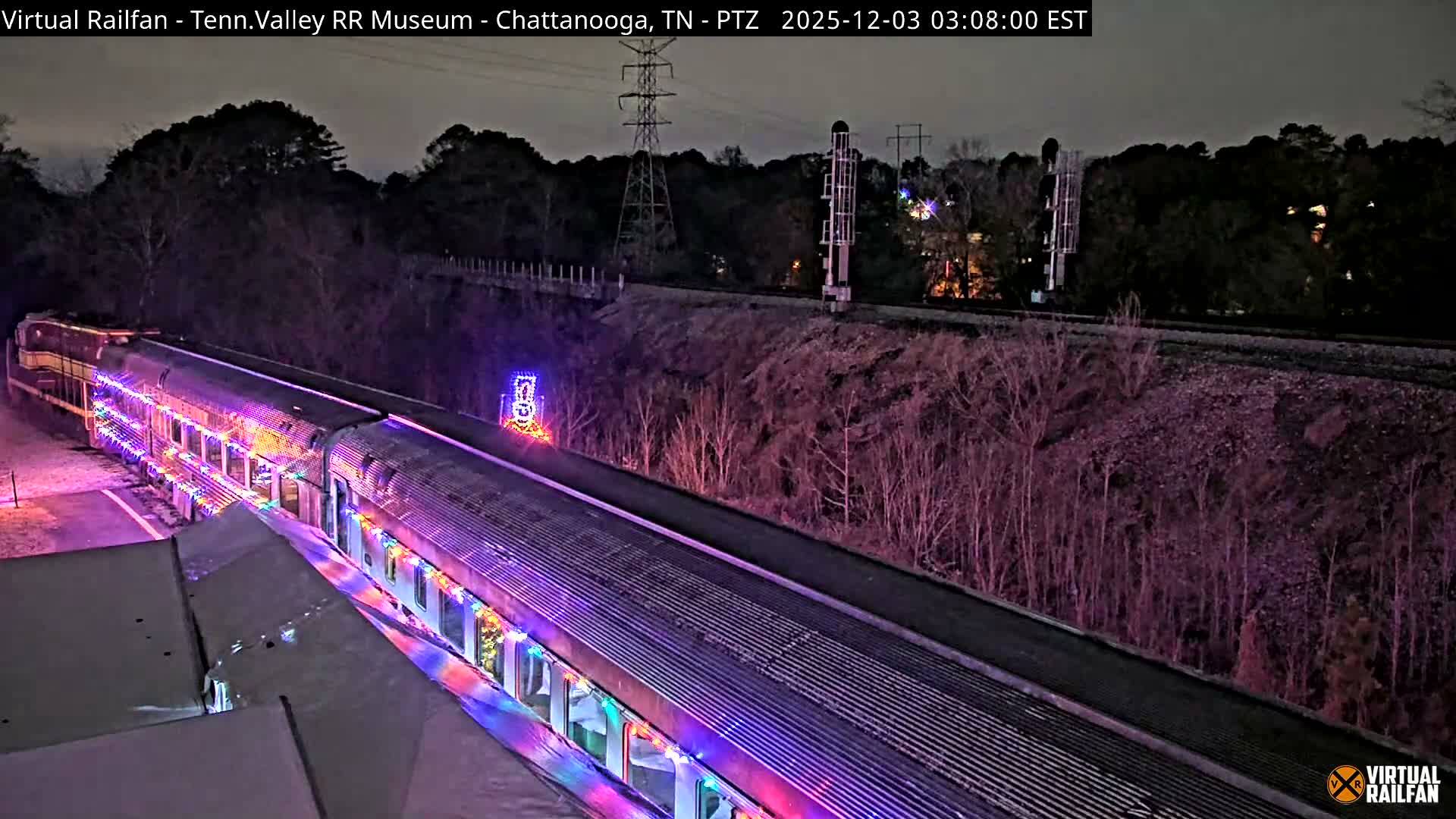 A long passenger train is brightly illuminated with colorful string lights while parked at night in an outdoor setting featuring bare trees and power lines under a clear, dark sky.