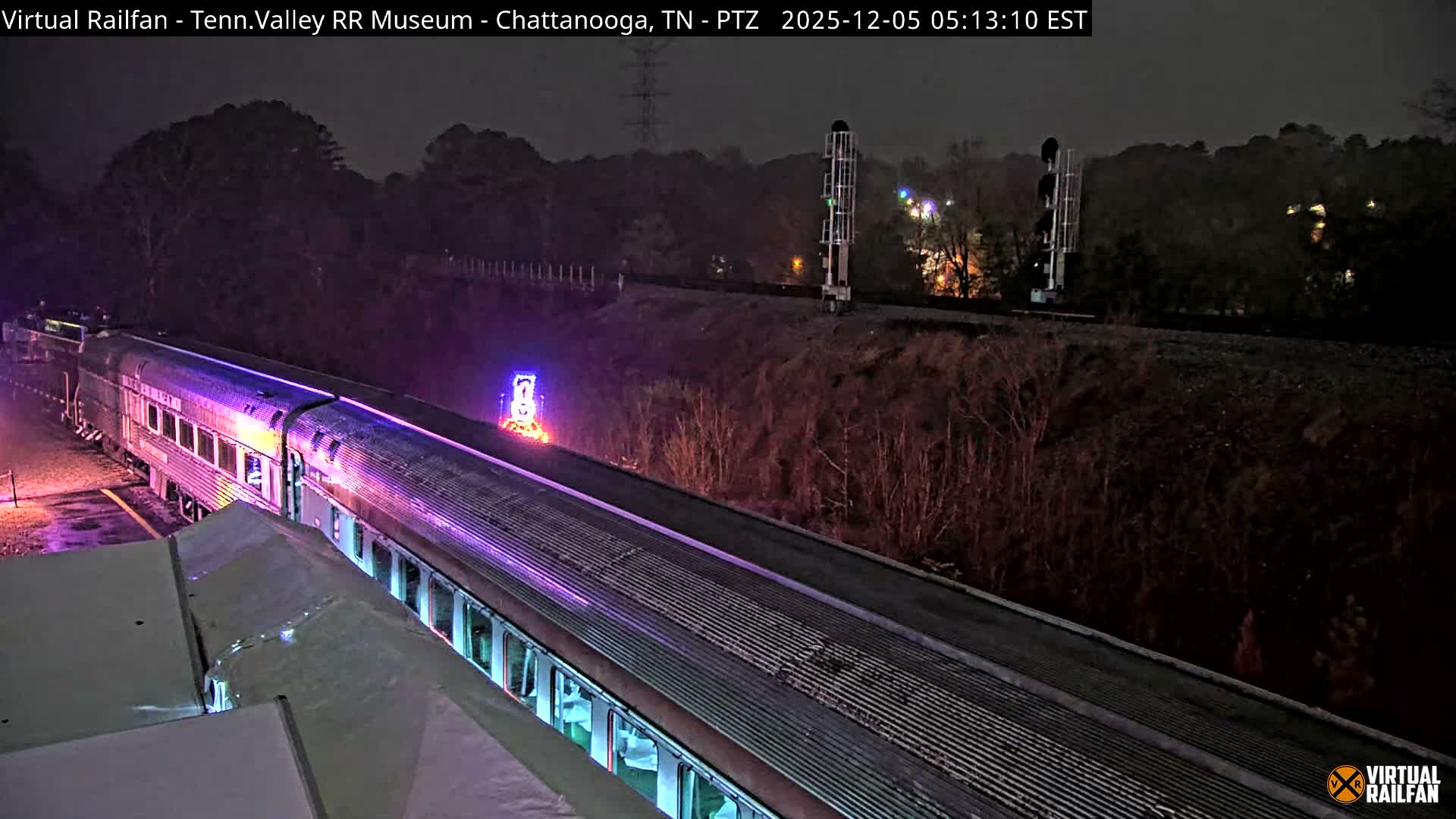 A long silver passenger train, illuminated with purple and other colorful lights, sits on tracks at night under an overcast sky with damp ground reflecting the lights, an illuminated "8" decoration nearby, and railroad signals visible further down the track.