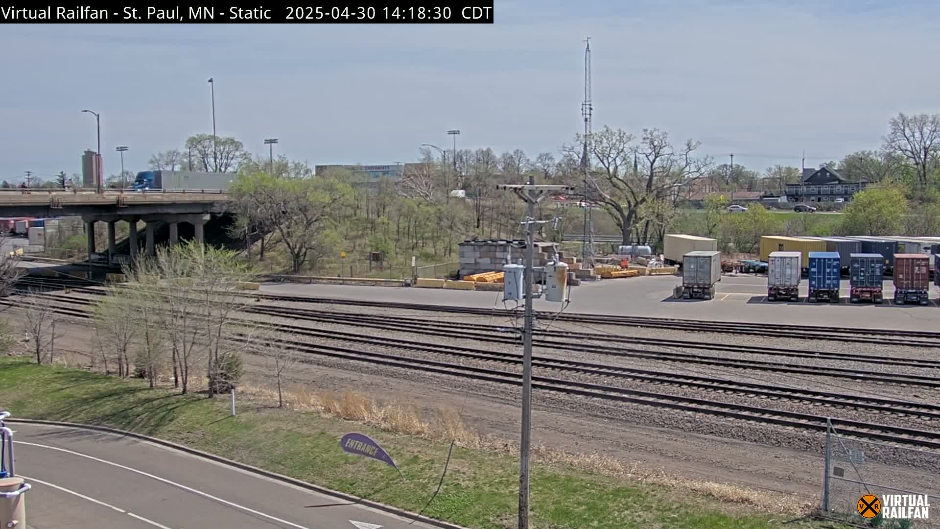 A sunny day reveals a railroad yard with several shipping containers, train tracks, a roadway, and a highway overpass, all surrounded by trees and vegetation.
