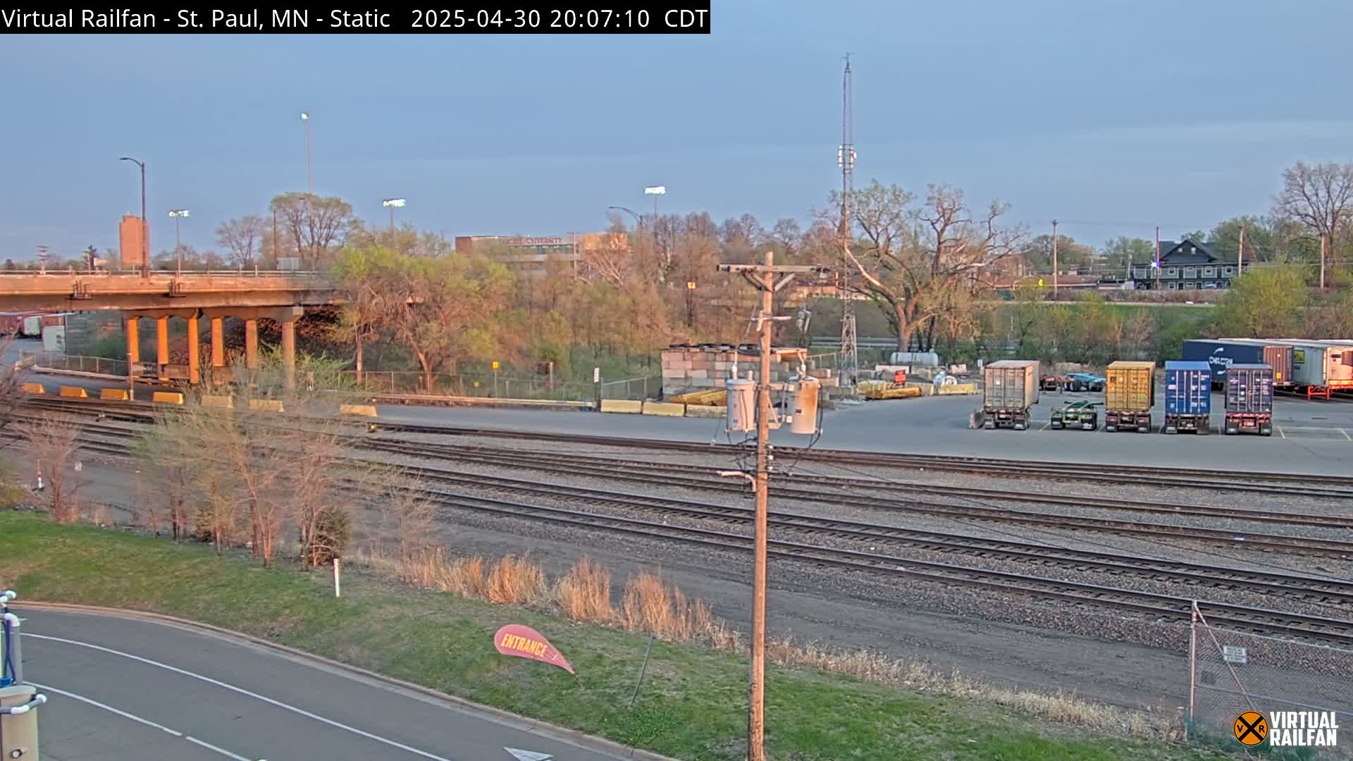Under a twilight sky, a railway yard with multiple train tracks, a road, and several shipping containers sits beside a low-lying overpass.