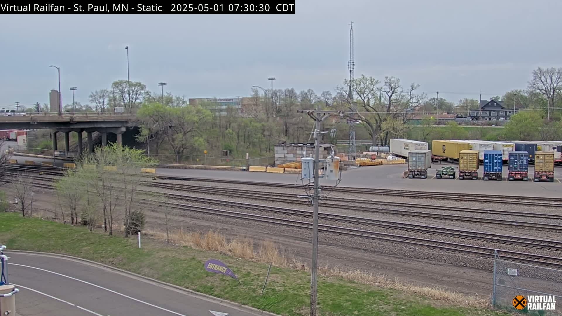 A cloudy day reveals a railway yard with multiple shipping containers, train tracks, a road, and a bridge overhead, surrounded by trees and greenery.